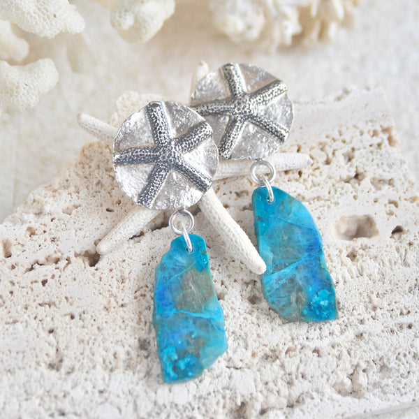 Pendientes "Nómada del Mar". Este diseño exclusivo combina la belleza natural de una estrella de mar con la piedra de crisocola en tonalidades que evocan las aguas cristalinas de una laguna secreta. En la parte superior, un medallón de plata de ley con un relieve de estrella de mar, La combinación de plata oxidada y plata blanca añade un contraste distintivo que realza la elegancia y el misterio del diseño. Almakaia by Heike Otten