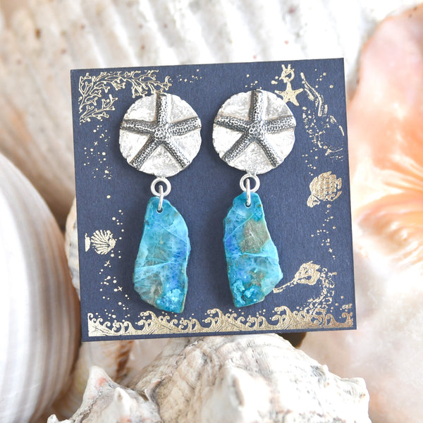 Pendientes "Nómada del Mar". Este diseño exclusivo combina la belleza natural de una estrella de mar con la piedra de crisocola en tonalidades que evocan las aguas cristalinas de una laguna secreta. En la parte superior, un medallón de plata de ley con un relieve de estrella de mar, La combinación de plata oxidada y plata blanca añade un contraste distintivo que realza la elegancia y el misterio del diseño. Almakaia by Heike Otten